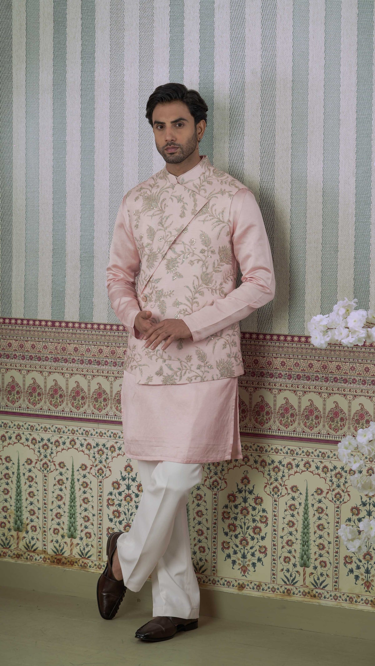 A man wearing a light pink embroidered bundi kurta with white trousers and dark brown shoes, standing against a patterned wallpaper background.