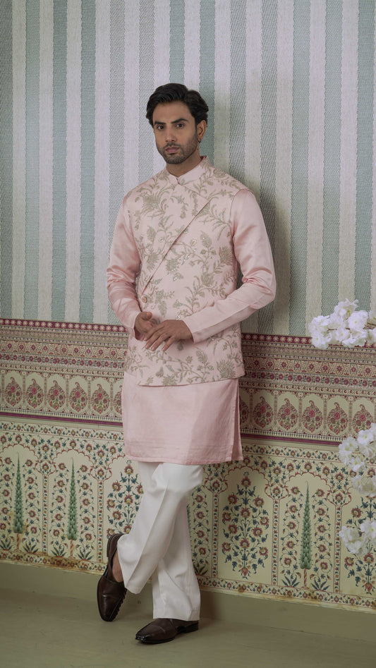 A man wearing a light pink embroidered bundi kurta with white trousers and dark brown shoes, standing against a patterned wallpaper background.