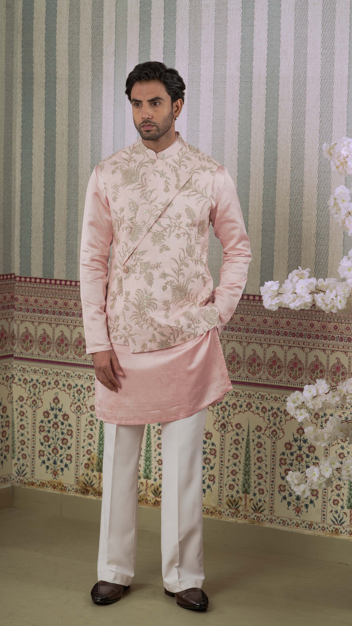 A man modeling a traditional Indian outfit consisting of a light pink bundi kurta with bluish floral embroidery and white trousers, standing in front of a patterned wallpaper background.