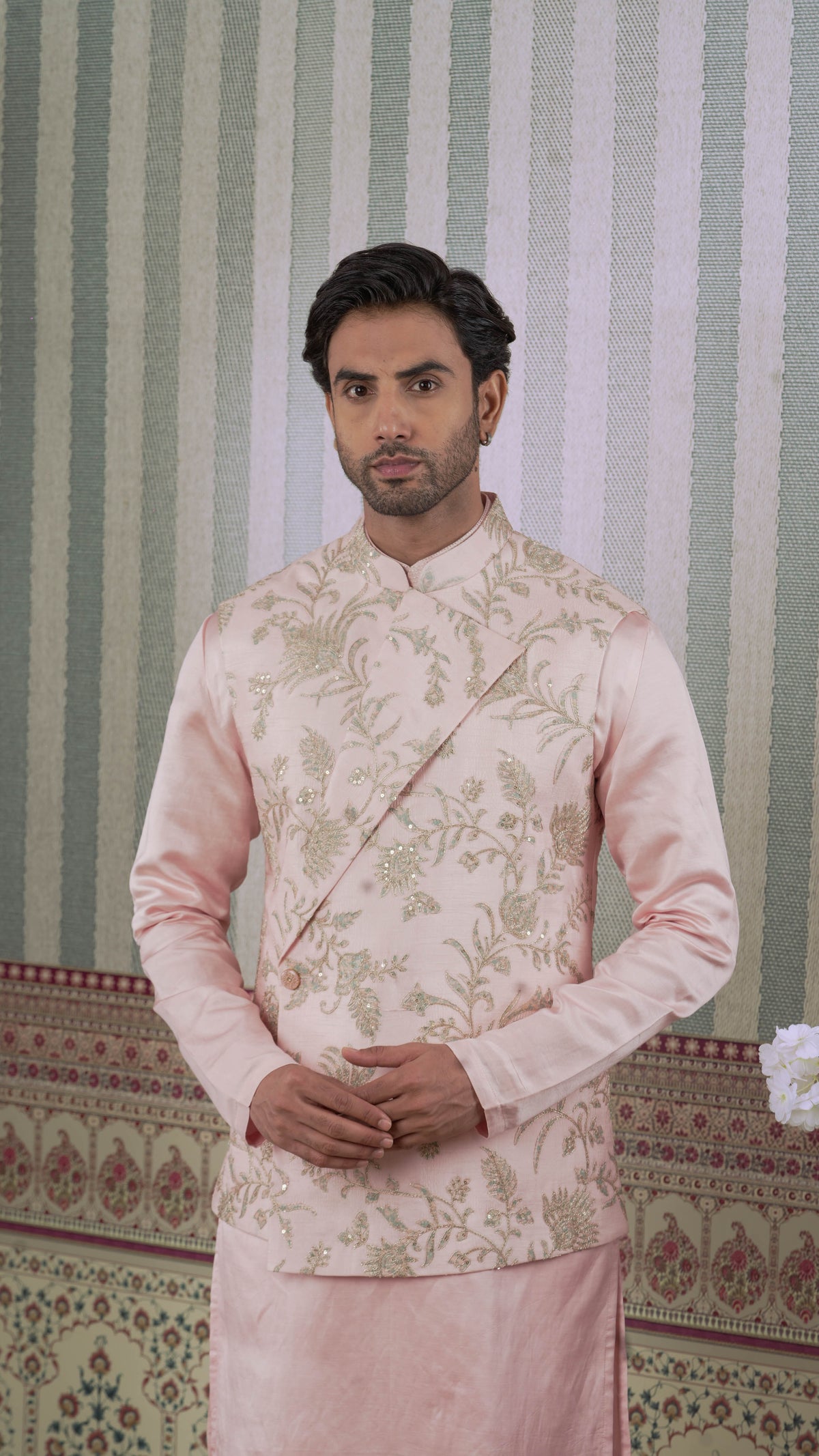A man wearing a light pink traditional Indian bundi jurta with bluish floral embroidery on the jacket, standing in front of a green and white striped wall.