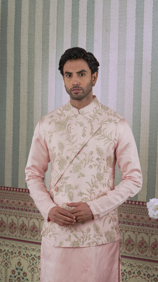 A man wearing a light pink traditional Indian bundi jurta with bluish floral embroidery on the jacket, standing in front of a green and white striped wall.