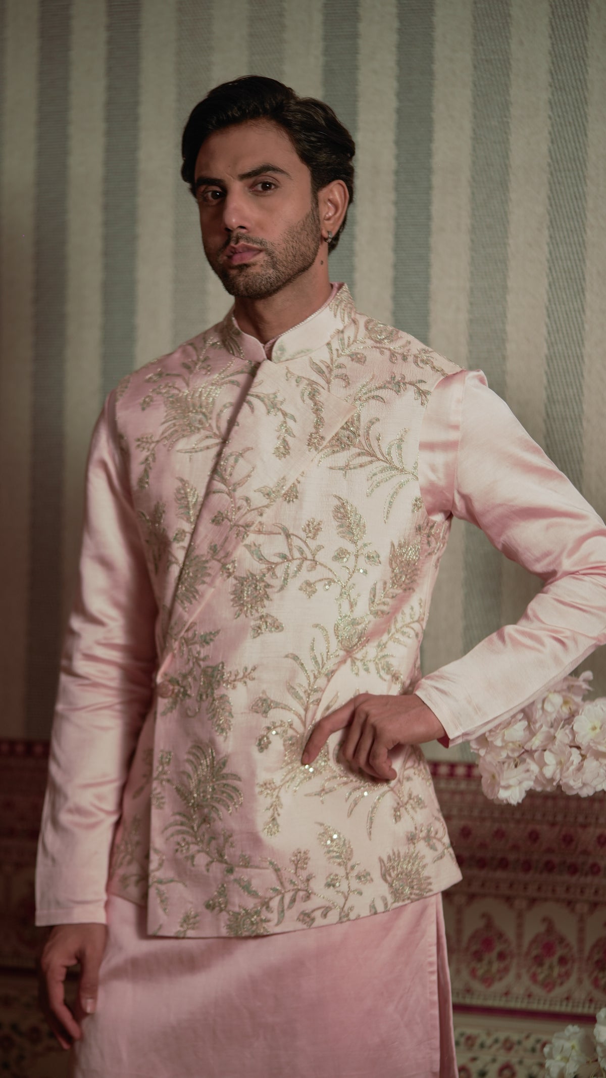 A man wearing a light pink bundi kurta Indian outfit with a heavily embroidered floral jacket and a matching pink kurta, standing against a striped wallpaper background.