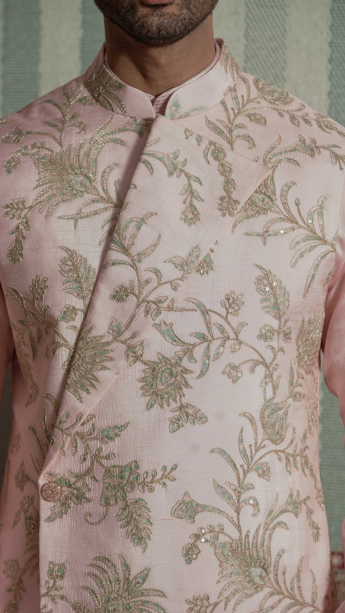 A close-up of a man wearing a light pink bundi jacket with intricate gold and bluish floral embroidery and sequin embellishments. The garment features a mandarin collar and a diagonal front closure.