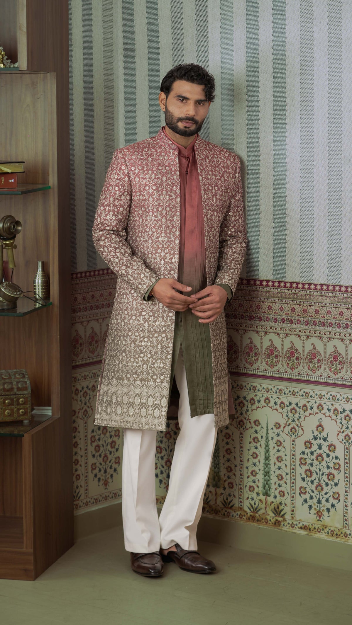 A man wearing a traditional Indian outfit stands in a decorated room. He is wearing a long, patterned sherwani jacket in ombre orange green, white trousers, and dark brown shoes.