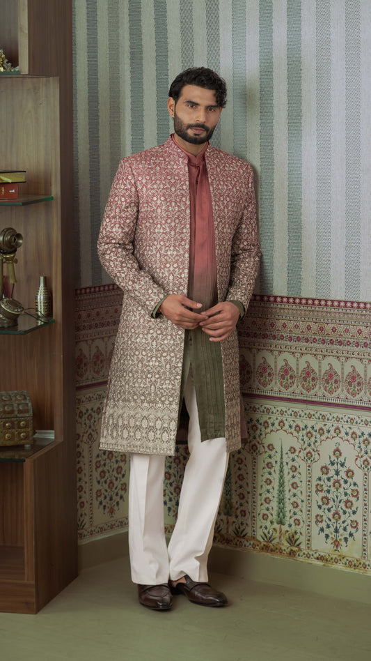 A man wearing a traditional Indian outfit stands in a decorated room. He is wearing a long, patterned sherwani jacket in ombre orange green, white trousers, and dark brown shoes.