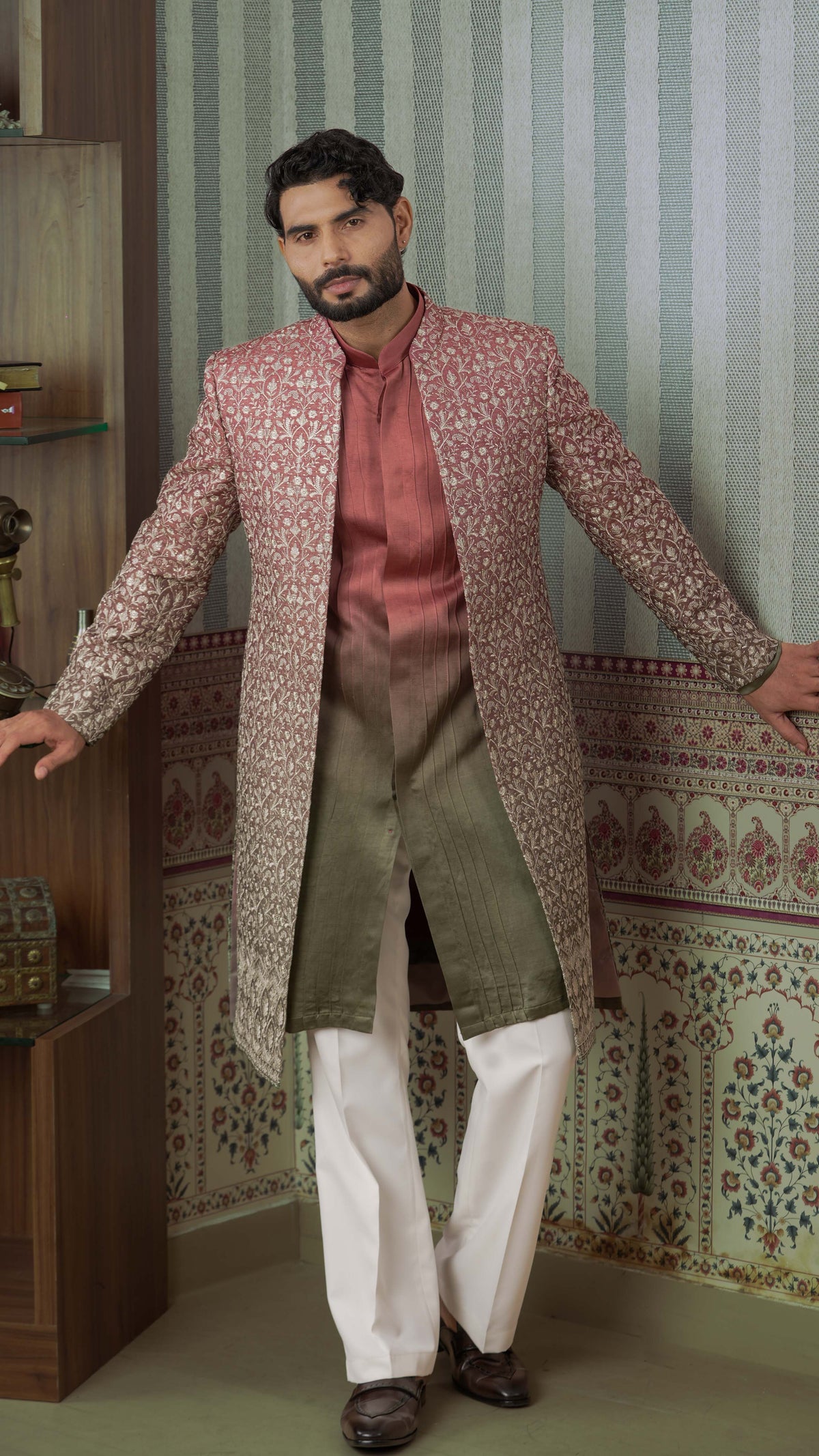 A man modeling a traditional Indian outfit featuring a long-sleeved, open-front sherwani jacket in a pink and gold floral pattern, worn over a rust-colored shirt, olive green kurta, white trousers, and dark brown shoes, posed against a patterned wallpaper background.