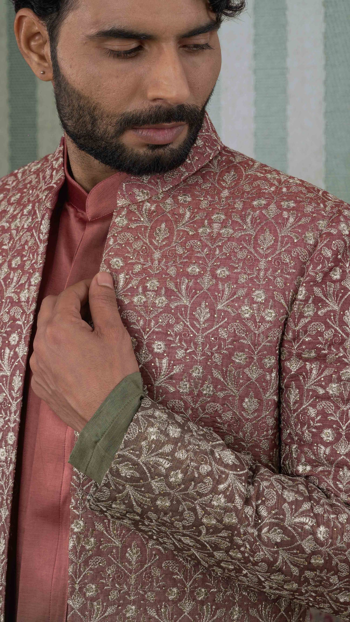 A close-up of a man wearing a orange green sherwani jacket with intricate gold floral embroidery, layered over a matching rose pink kurta and a green cuff.