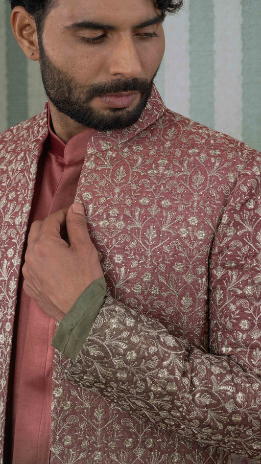 A close-up of a man wearing a orange green sherwani jacket with intricate gold floral embroidery, layered over a matching rose pink kurta and a green cuff.
