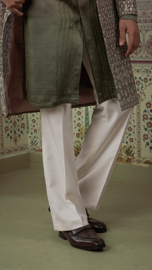 A person wearing traditional featuring an orange olive green kurta, a brown patterned overcoat, off-white trousers, and dark brown leather loafers, standing against a patterned wall.