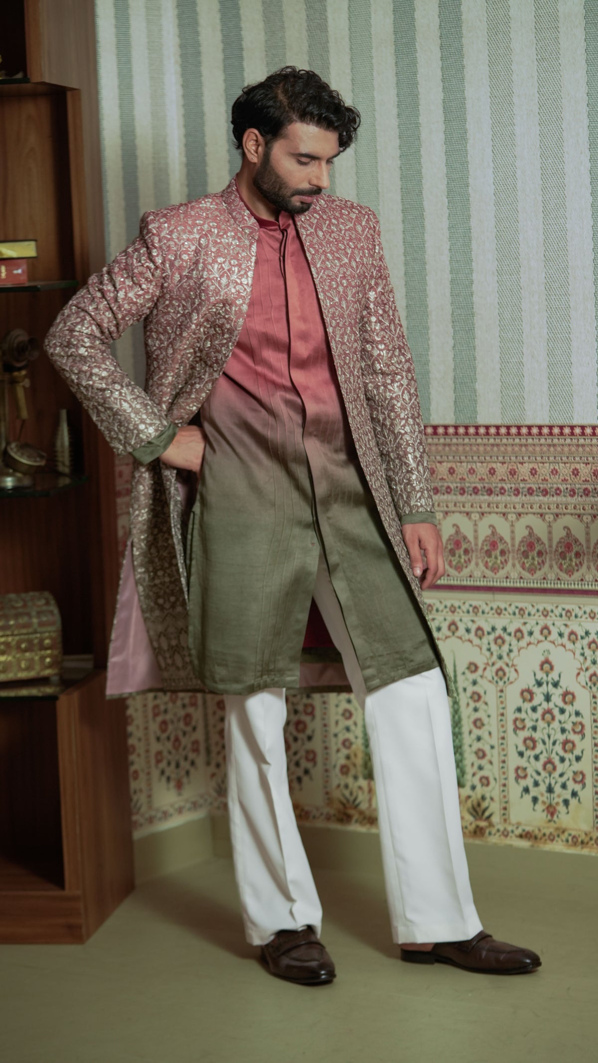 A man modeling a traditional Sherwani Kurta outfit featuring an ombre metallic gold patterned sherwani over a dusty rose kurta, an olive green drape, white trousers, and brown leather loafers, set against a patterned wallpaper background