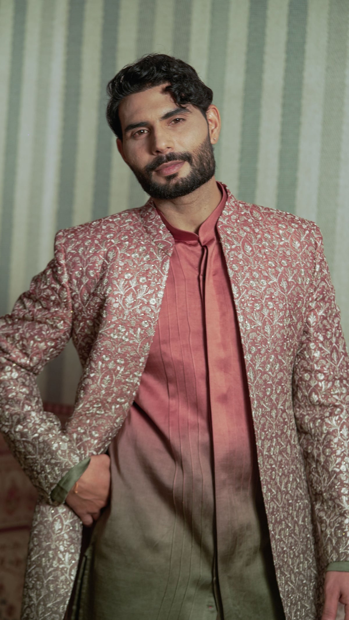 A man wearing a metallic rose gold and gold patterned sherwani jacket over a matching rose gold kurta and olive green trousers, posed against a green and white striped background.