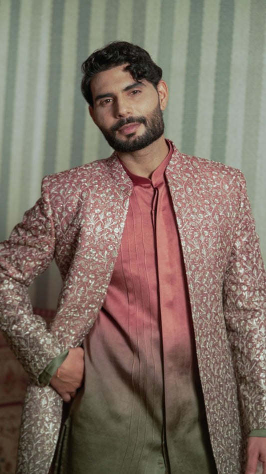 A man wearing a metallic rose gold and gold patterned sherwani jacket over a matching rose gold kurta and olive green trousers, posed against a green and white striped background.