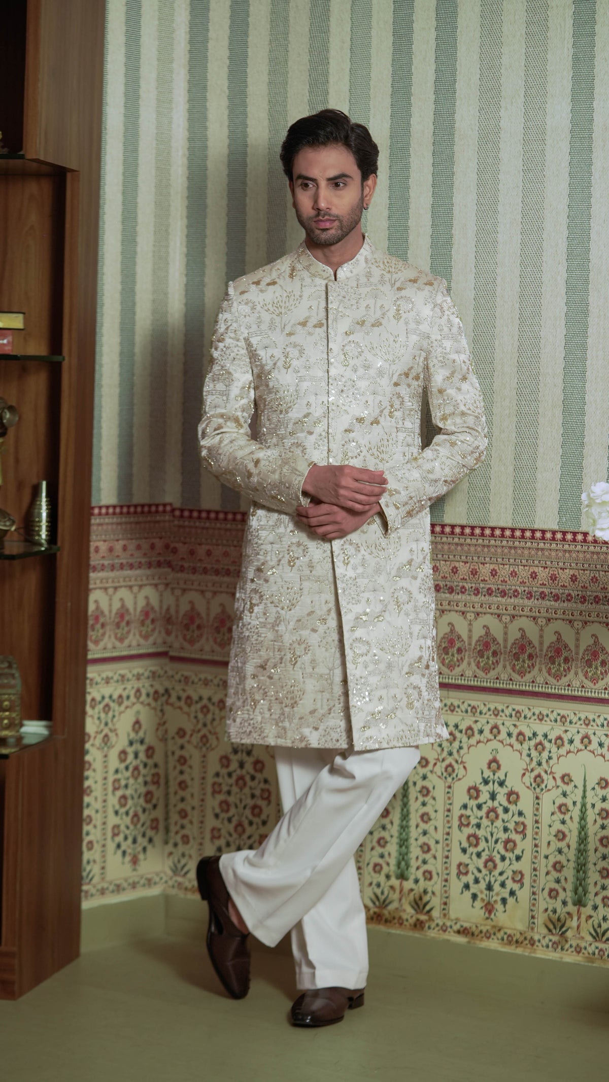 A man wearing a cream-colored, heavily embroidered sherwani jacket with white trousers, standing in front of a patterned wallpaper background.