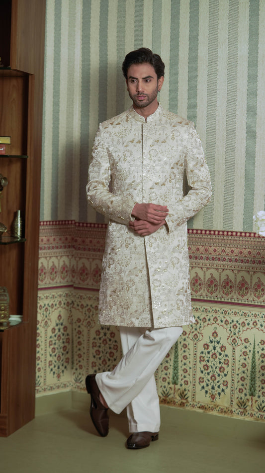 A man wearing a cream-colored, heavily embroidered sherwani jacket with white trousers, standing in front of a patterned wallpaper background.