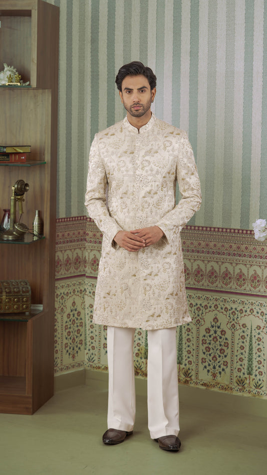 A man wearing a beige-colored embroidered sherwani jacket with white trousers, standing in front of a patterned green and white striped wall.