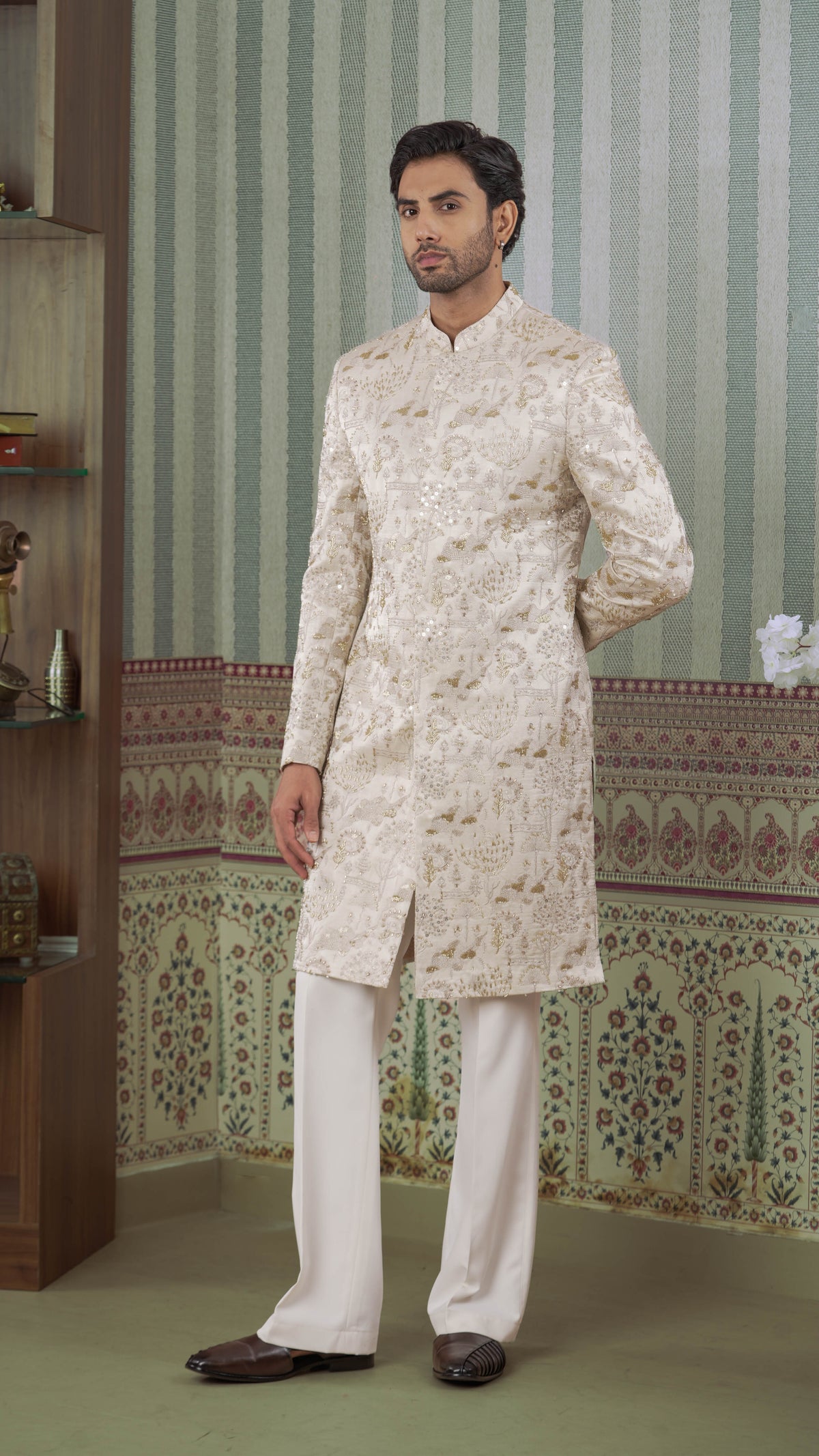 A man modeling a traditional Indian outfit consisting of a beige-colored embroidered sherwani jacket and white trousers, standing in front of a patterned wallpaper background.