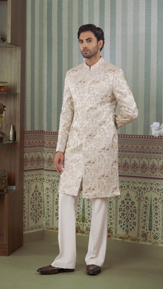 A man modeling a traditional Indian outfit consisting of a beige-colored embroidered sherwani jacket and white trousers, standing in front of a patterned wallpaper background.