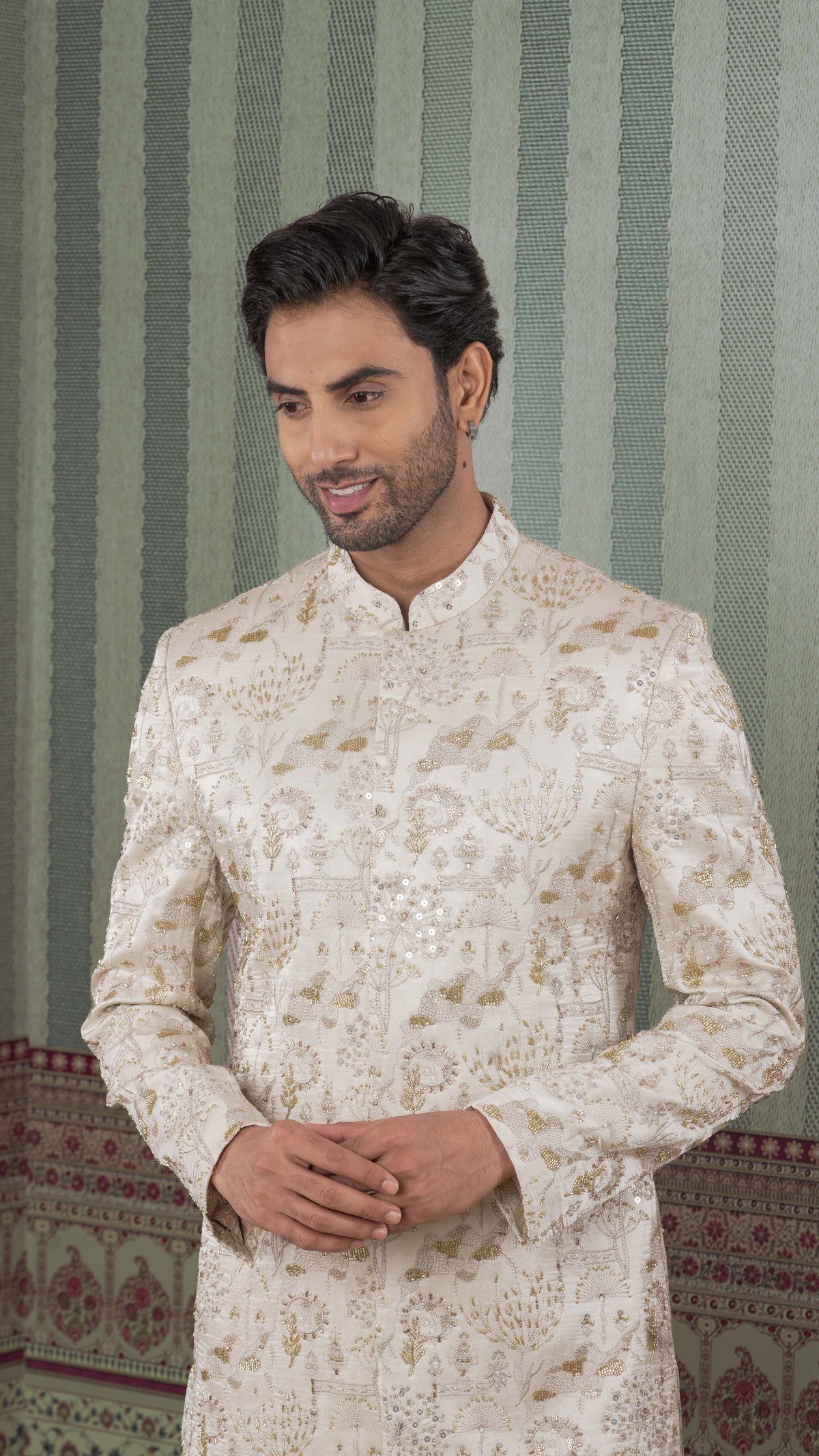 A man wearing a beige-colored sherwani jacket with intricate gold floral embroidery, standing in front of a green and white striped patterned wall.