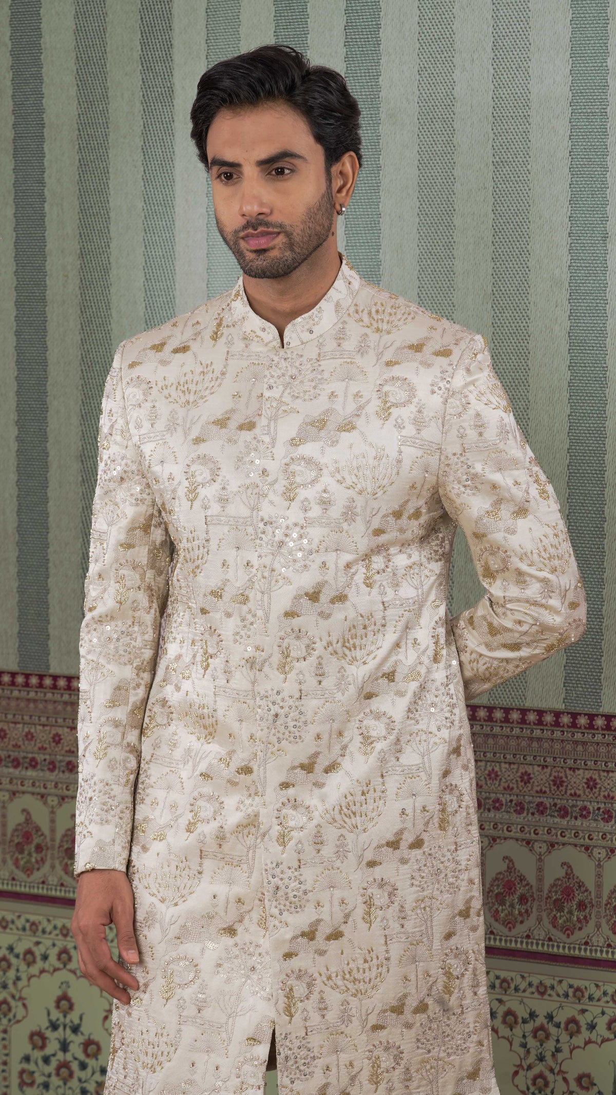 A man wearing a beige-colored, long-sleeved sherwani jacket with intricate gold floral embroidery, standing against a patterned green and beige striped wall.