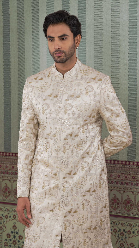 A man wearing a beige-colored, long-sleeved sherwani jacket with intricate gold floral embroidery, standing against a patterned green and beige striped wall.
