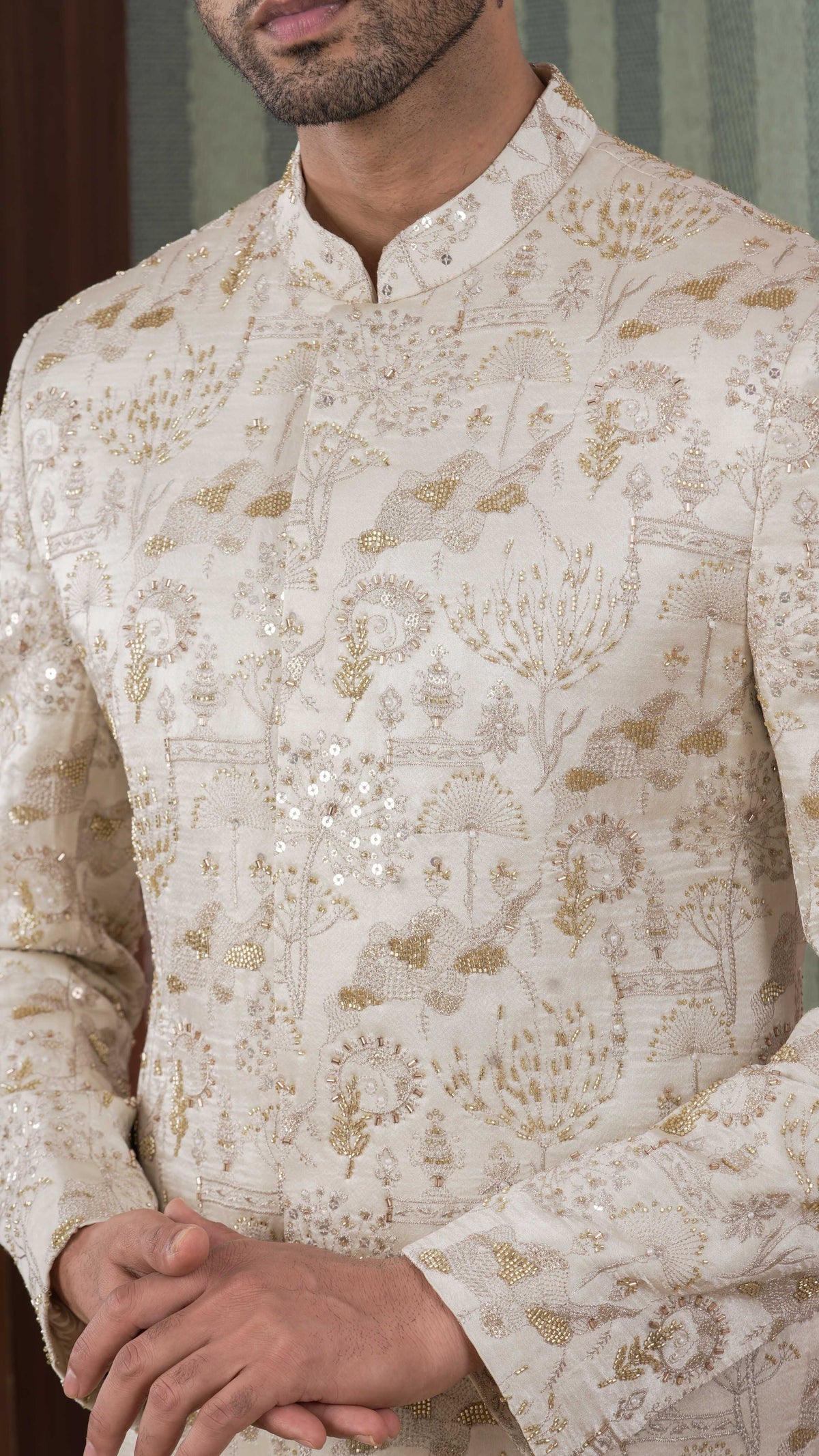 A close-up of a man wearing a beige-colored sherwani jacket with intricate gold and silver embroidery and mirror work, featuring a mandarin collar and long sleeves.
