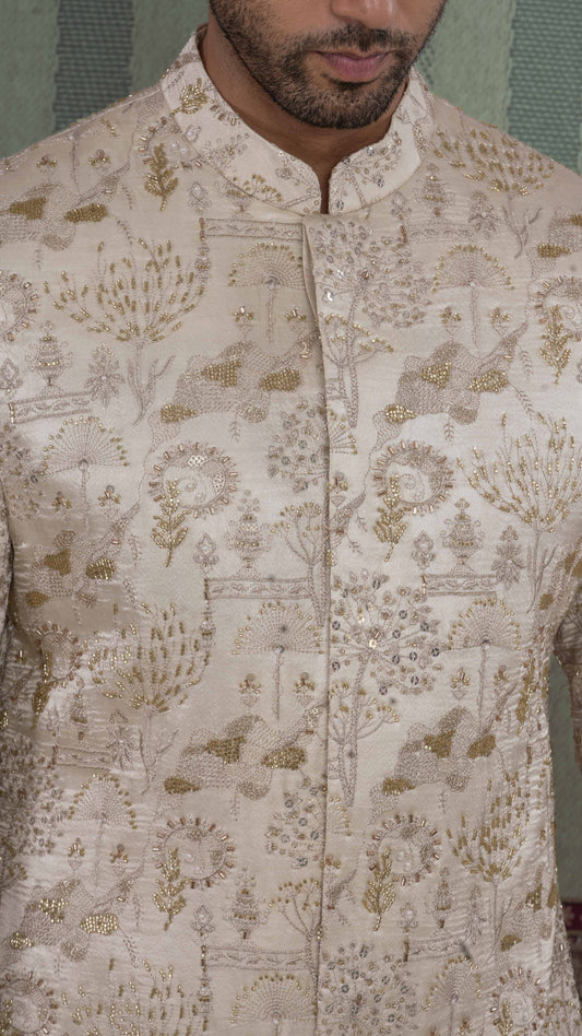 A close-up of a man wearing a beige-colored sherwani jacket with intricate gold and silver embroidery and mirror work, featuring a mandarin collar and buttoned front.