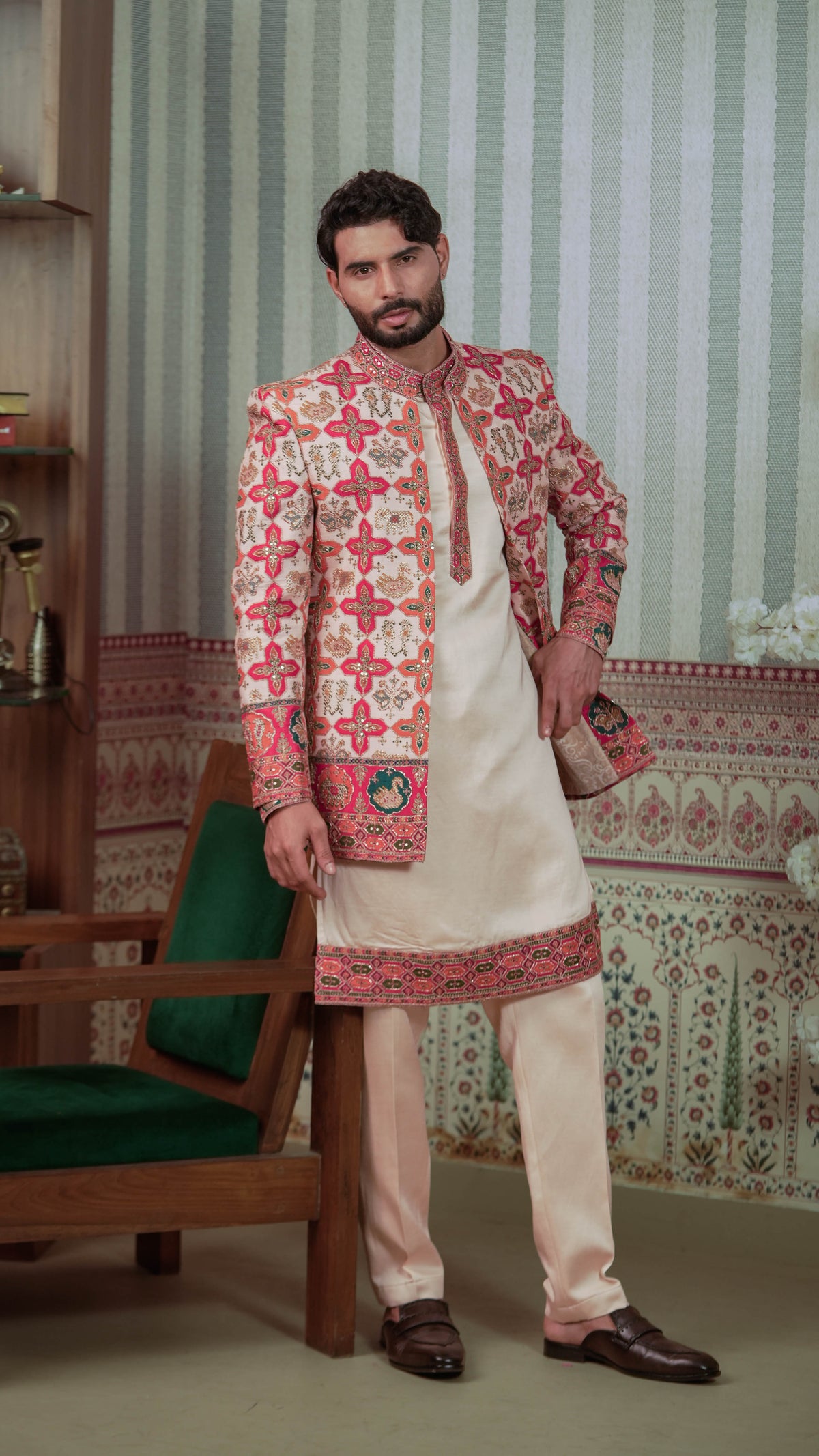 A man modeling a traditional Indian outfit consisting of a cream kurta with a heavily embroidered red, pink, and gold patterned jacket, cream trousers, and brown leather loafers, standing in a decorated room with patterned wallpaper and a green chair.