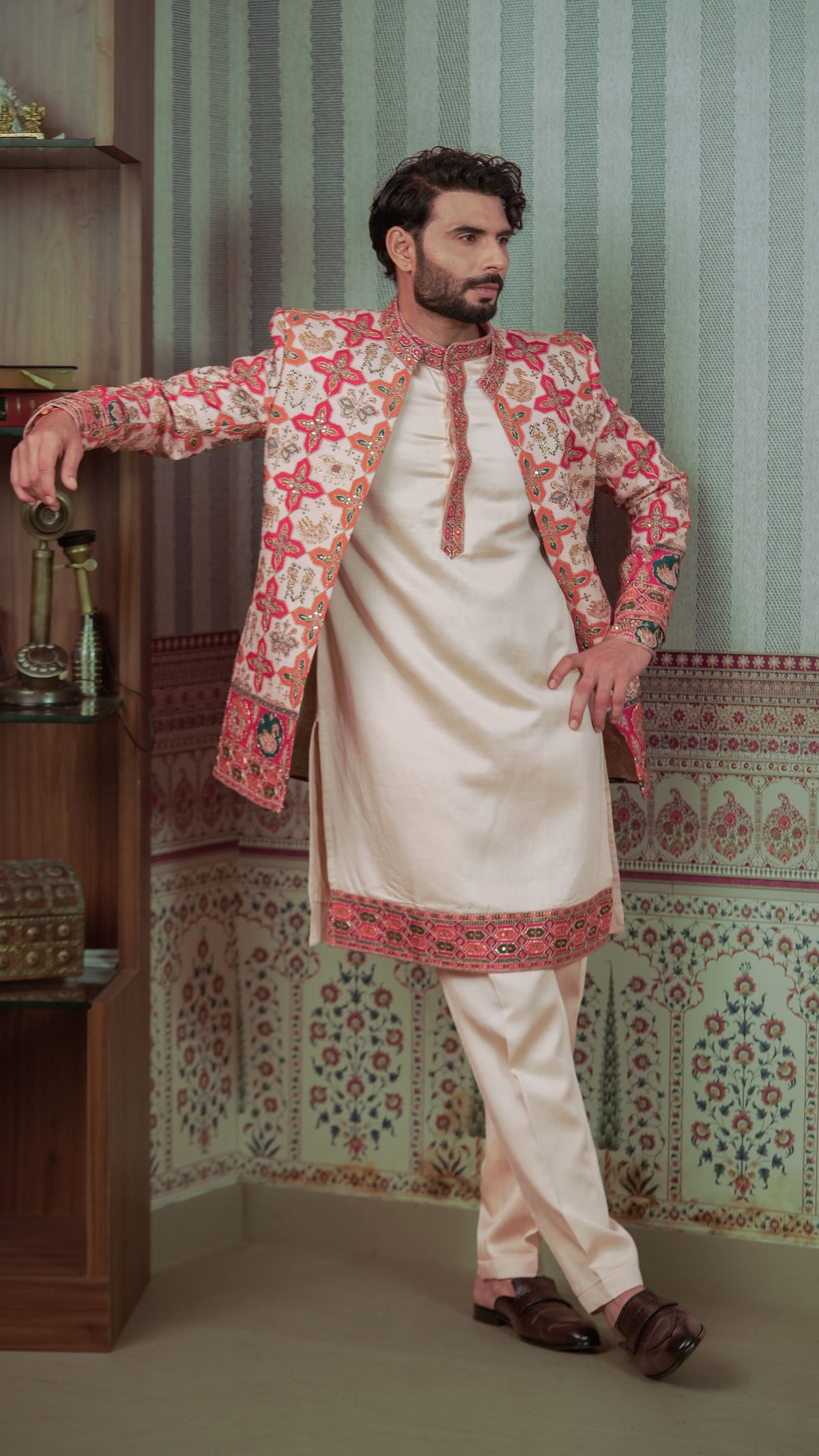 A man models a traditional Patola Jodhpuri consisting of a cream-colored kurta and trousers with a matching embroidered jacket. The jacket features intricate red, pink, and gold floral patterns.