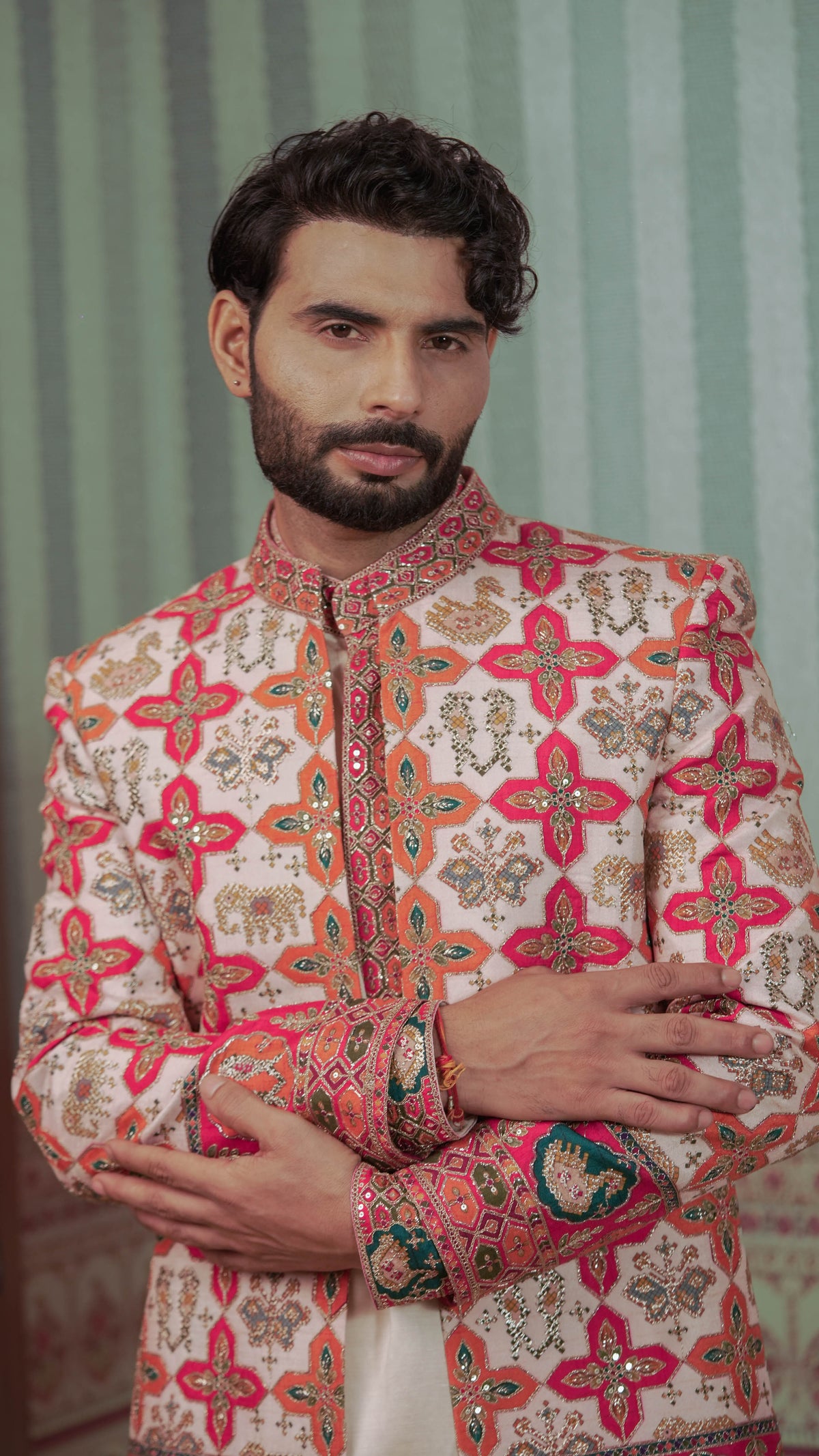 A man wearing a traditional Indian sherwani jacket with intricate multicolor embroidery and a mandarin collar, posed against a green striped background.