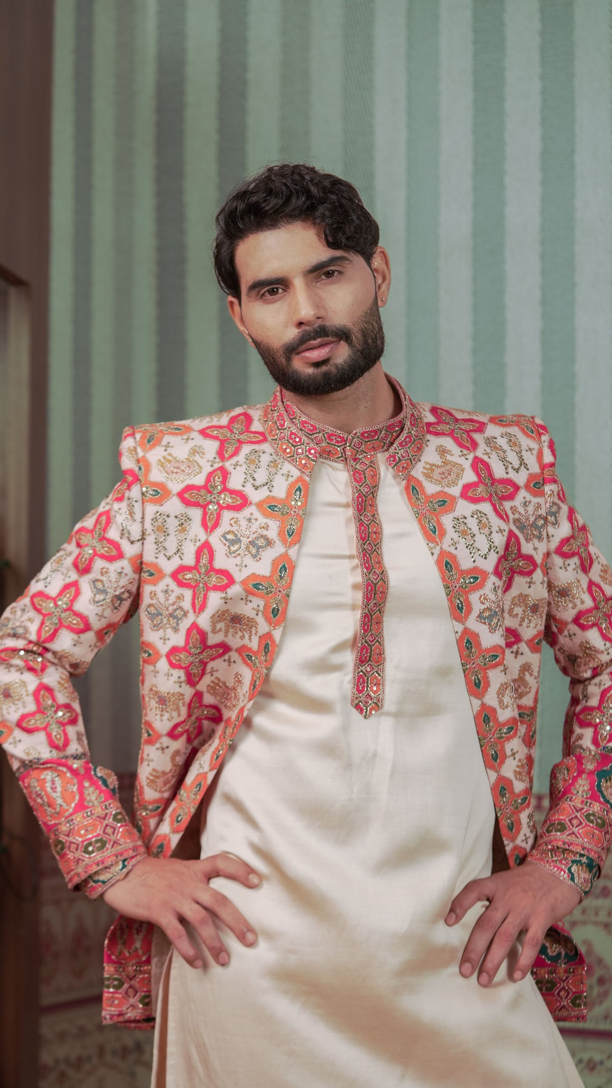A man wearing a cream-colored kurta with a heavily embroidered peach, pink, and red patterned sherwani jacket, posed against a green striped background.