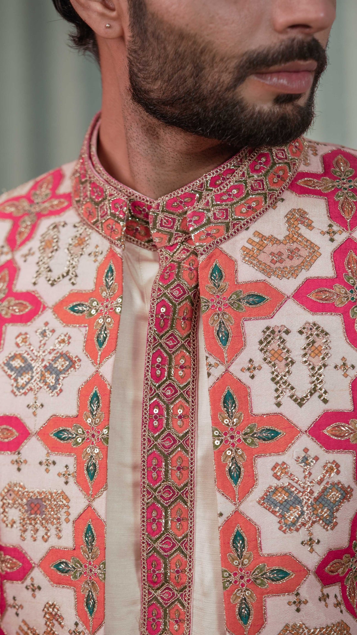 Close-up of a man wearing a cream-colored traditional sherwani jacket with vibrant pink, orange, and green geometric embroidery and metallic gold thread detailing.