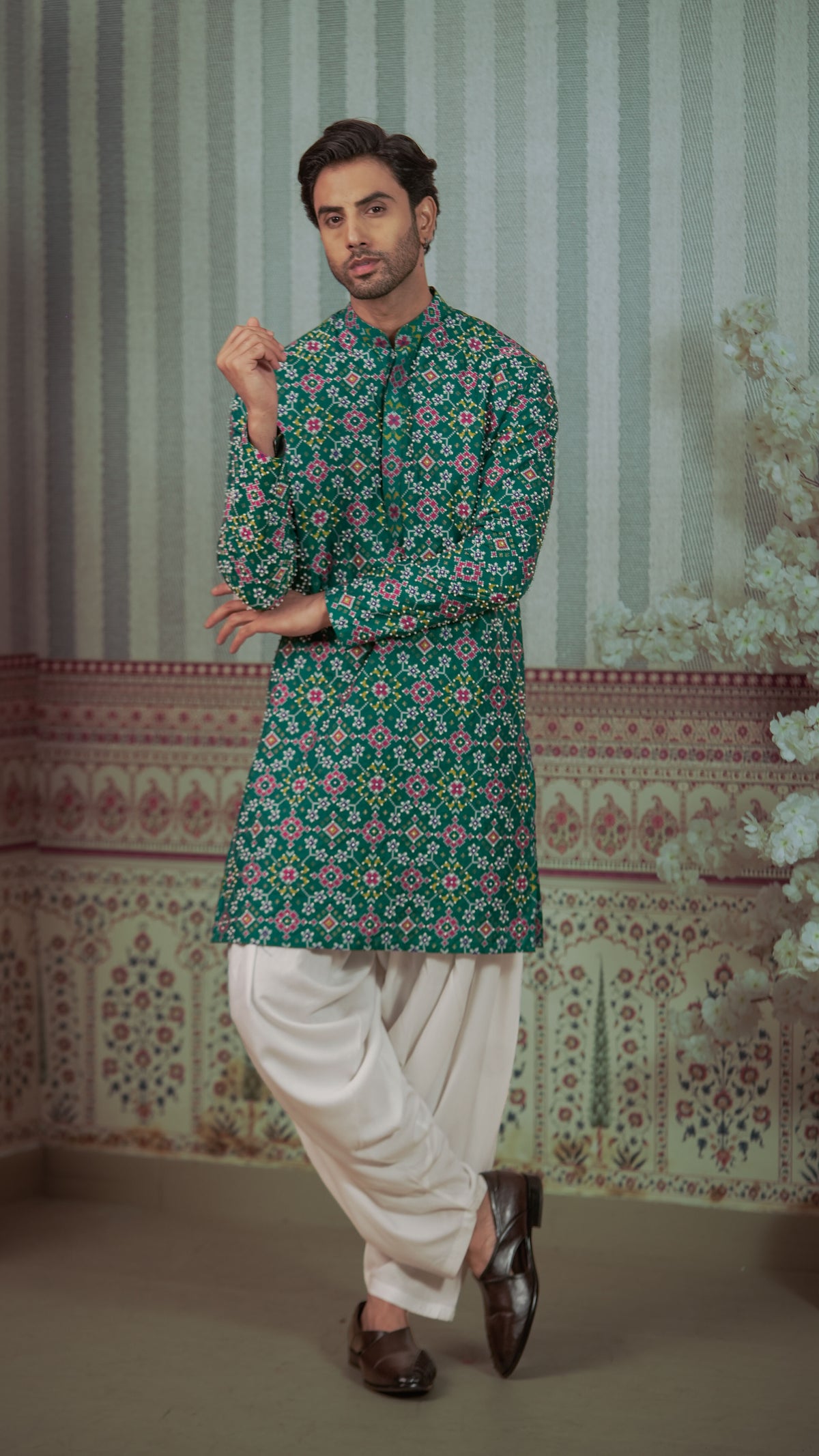 A man modeling a green long-sleeve kurta with a pink and white floral pattern, paired with white pants and dark brown shoes, standing in front of a patterned wall backdrop.