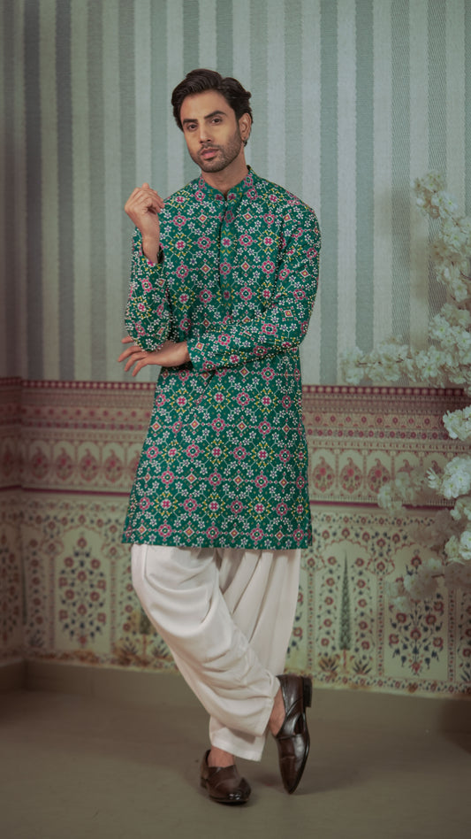 A man modeling a green long-sleeve kurta with a pink and white floral pattern, paired with white pants and dark brown shoes, standing in front of a patterned wall backdrop.