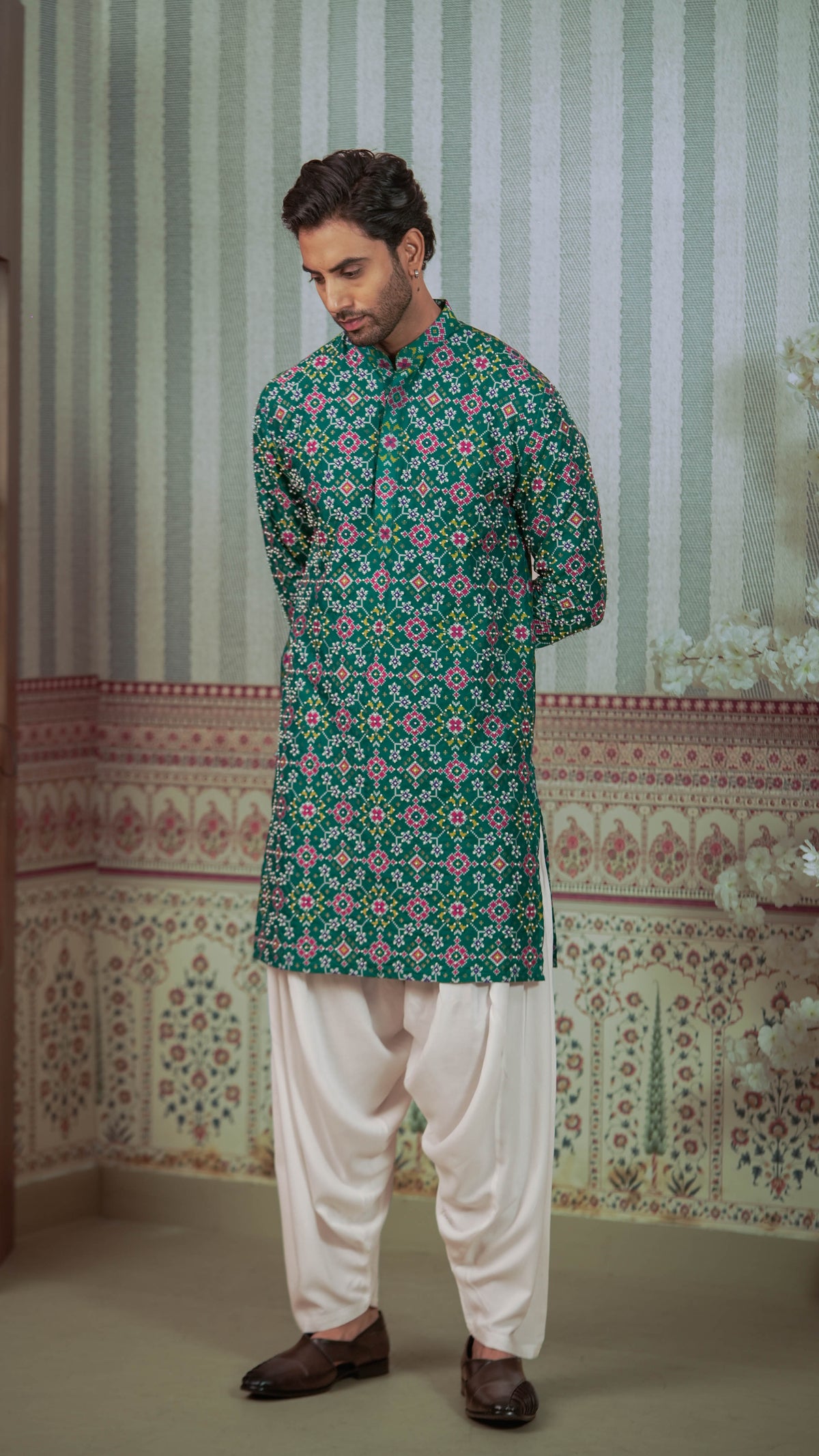 A man wearing a green patterned kurta with white pants and brown shoes, standing in front of a patterned wall.