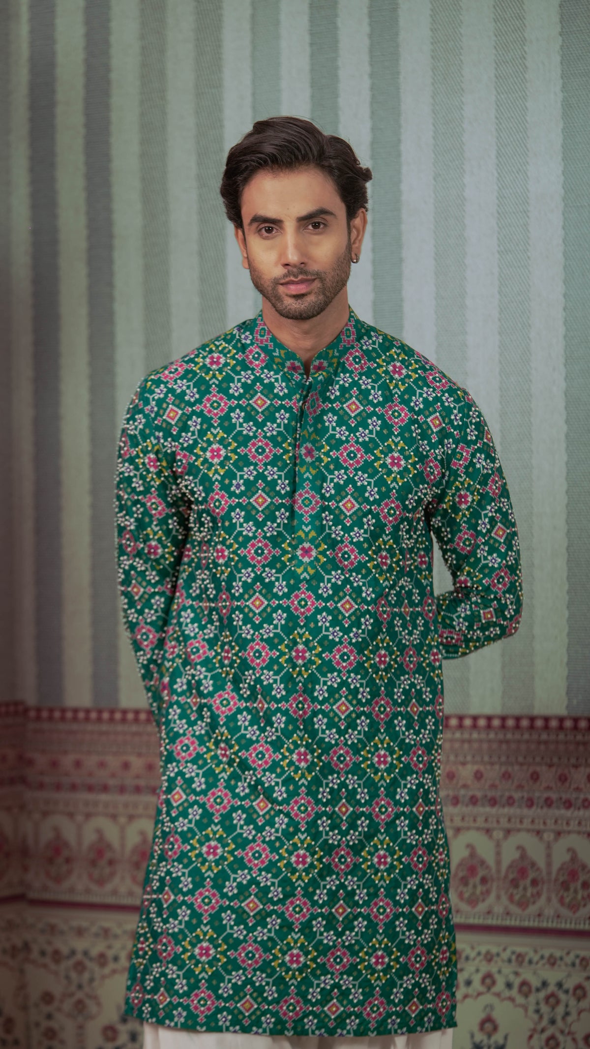 A man wearing a green long-sleeve kurta with a colorful geometric floral pattern, standing in front of a patterned green and white striped backdrop.