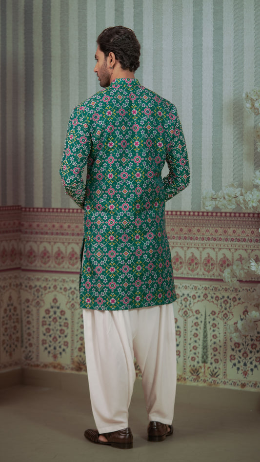 A man viewed from behind wearing a green patola kurta with white patiala and brown shoes, standing in front of a patterned indoor backdrop.
