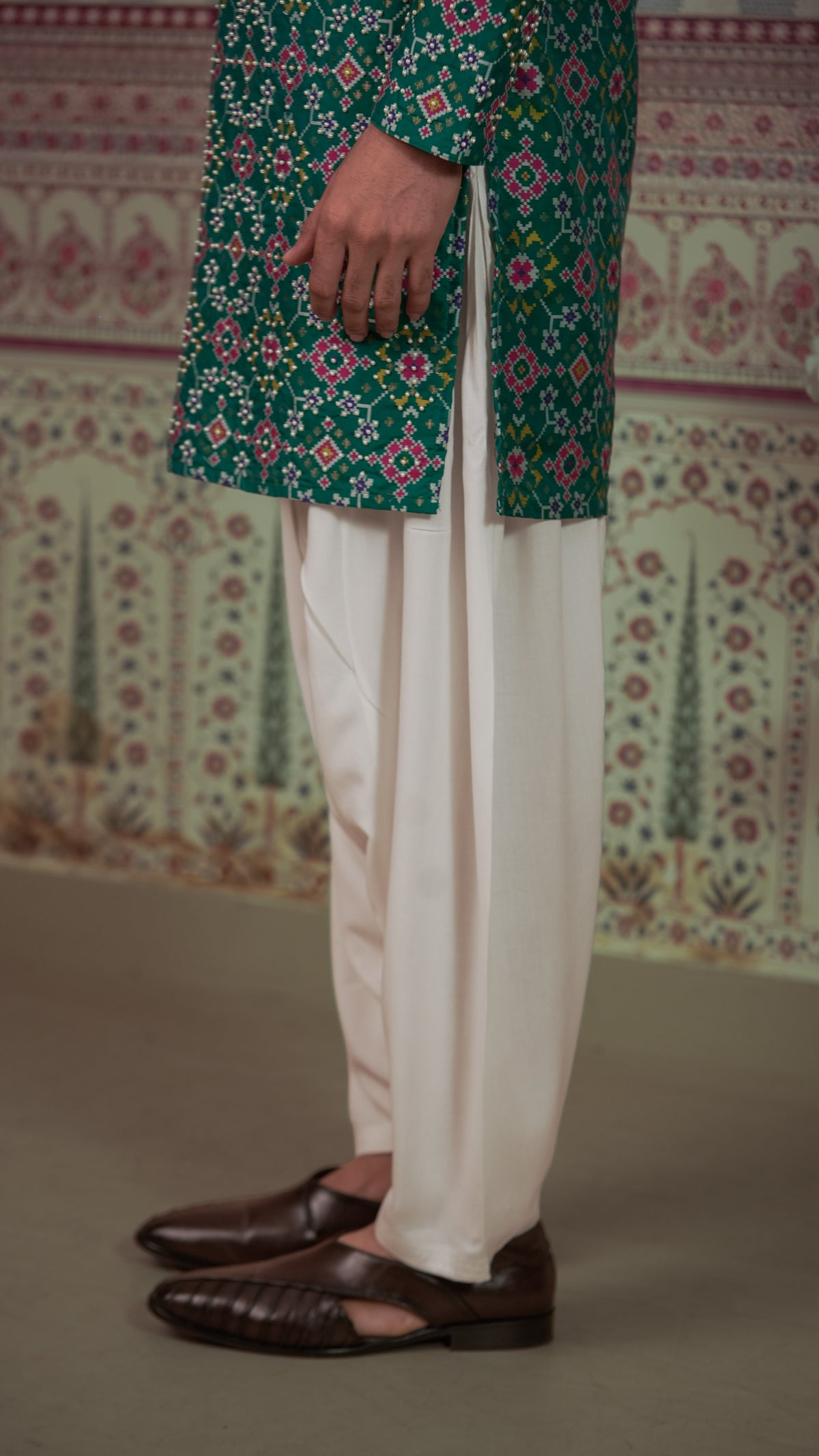 A person wearing a green patola kurta with white patiala and dark brown leather loafers, standing in front of a patterned wall.