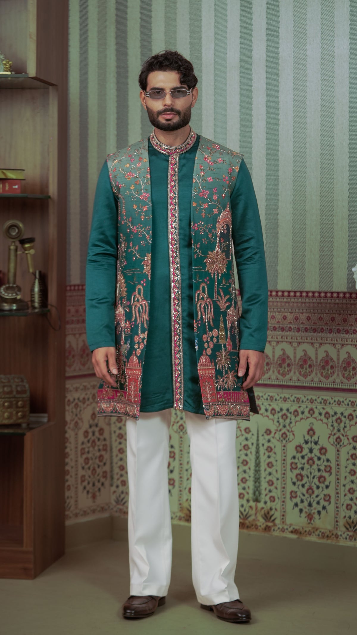 A man wearing a traditional Indian outfit consisting of a teal green kurta with intricate multicolor embroidery, white trousers, and brown shoes, standing in front of a patterned wall.