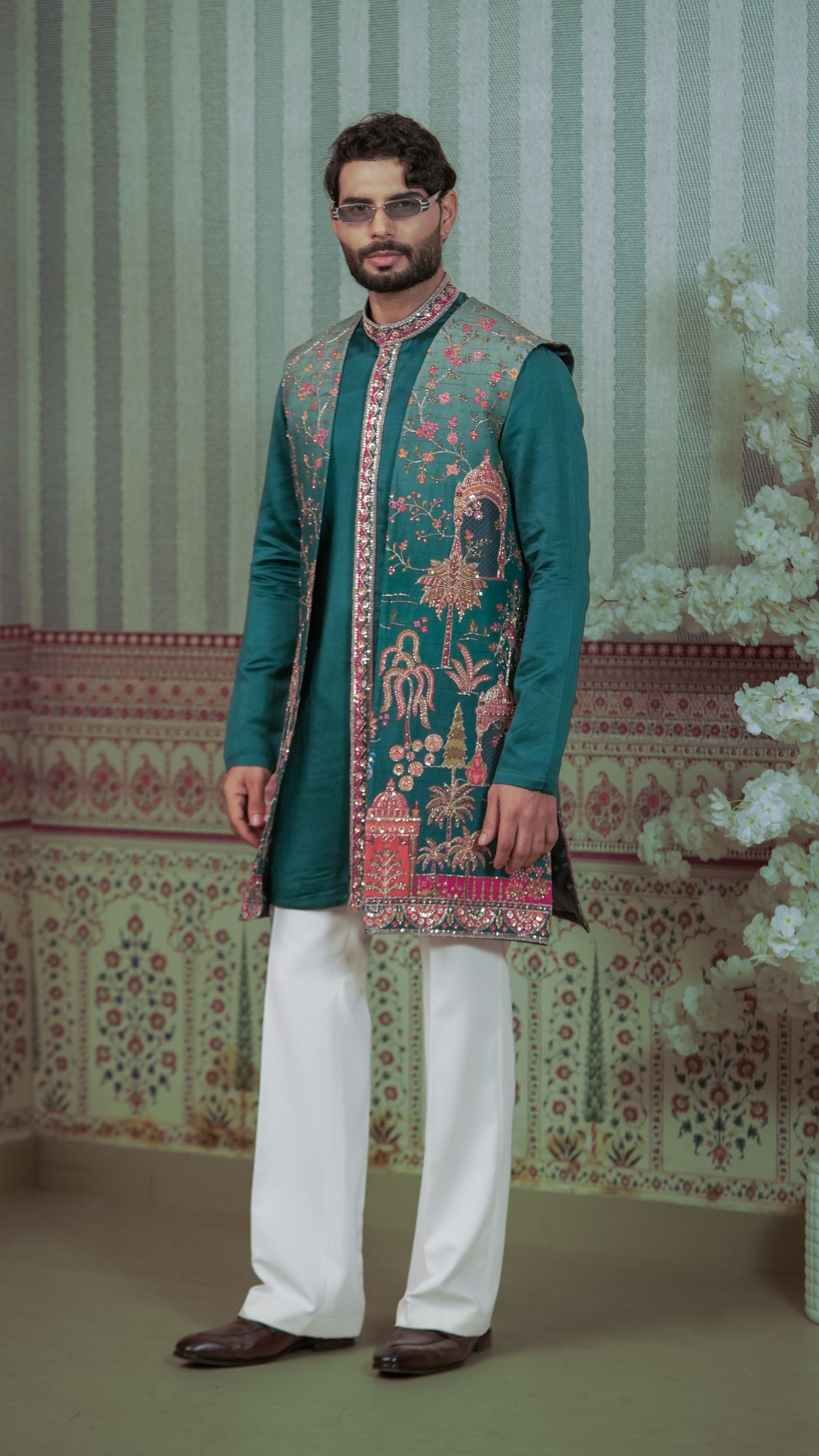 A man wearing a traditional Indian outfit consisting of a teal green kurta with intricate pink and gold embroidery, white trousers, and brown shoes, standing in front of a patterned green and white backdrop.