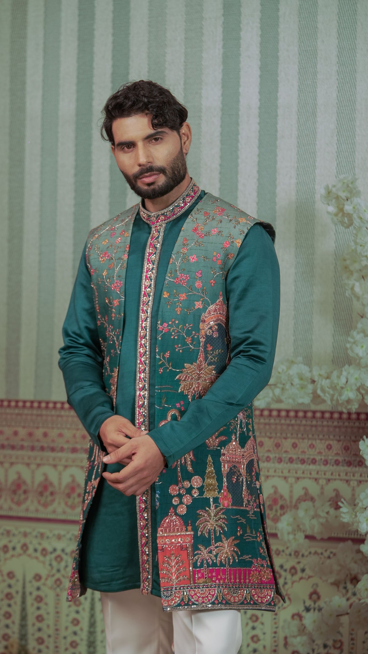 A man wearing a dark teal green embroidered kurta with a matching embroidered vest, paired with white trousers, standing against a patterned green and white background.
