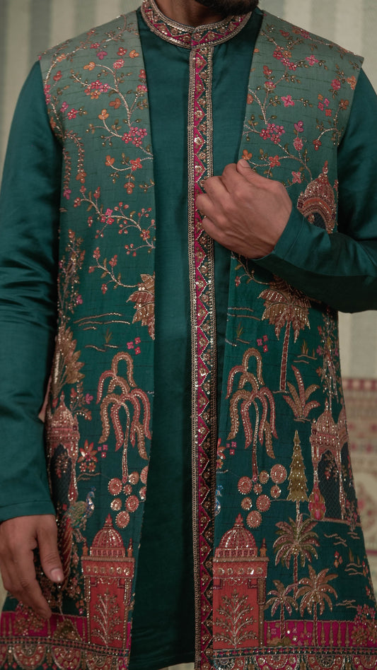 A close-up of a person wearing a dark teal green embroidered sherwani jacket with intricate gold, pink, and red floral and architectural motifs, paired with a matching green kurta.