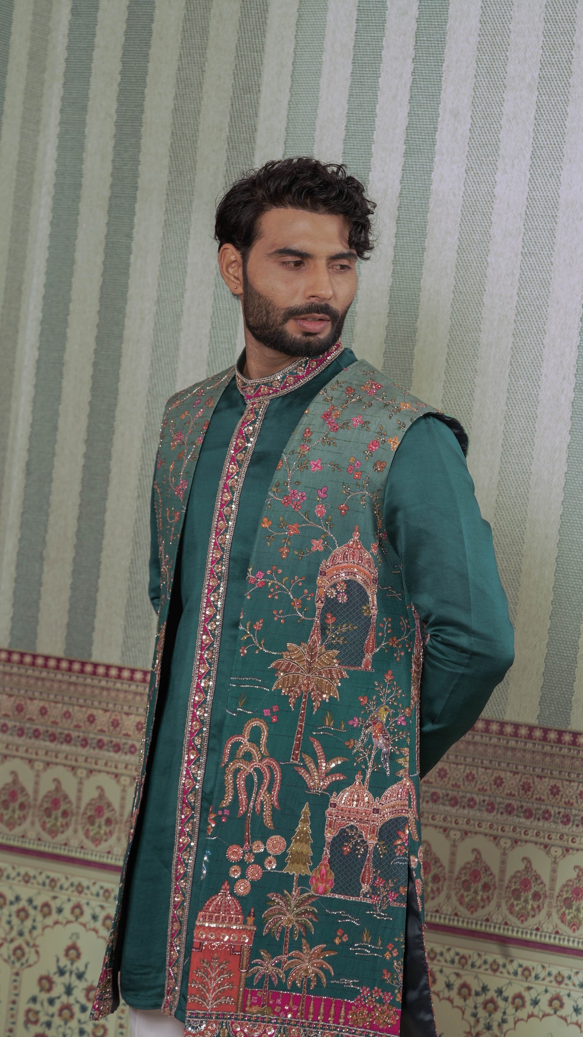 A man wearing a dark teal green traditional kurta with ornate pink and gold embroidery, standing against a green and white striped wallpaper background.