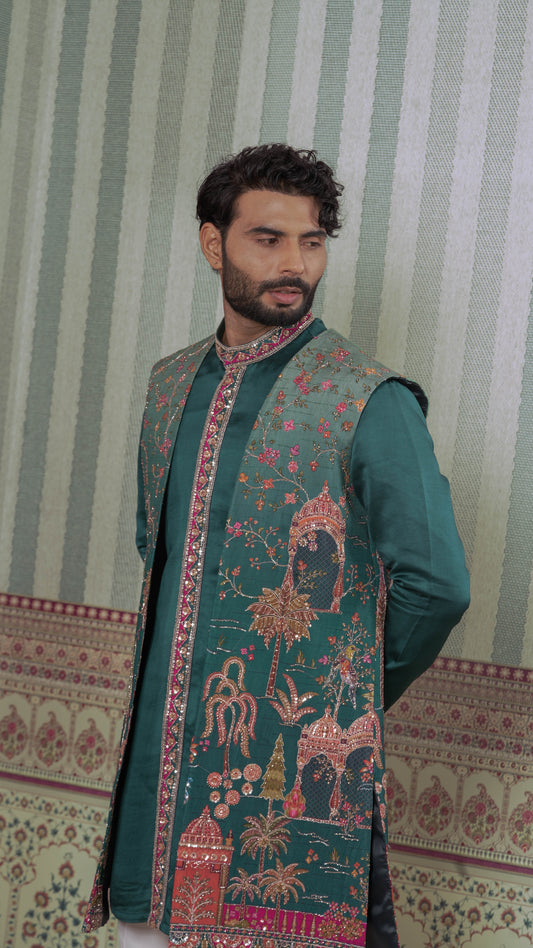 A man wearing a dark teal green traditional kurta with ornate pink and gold embroidery, standing against a green and white striped wallpaper background.