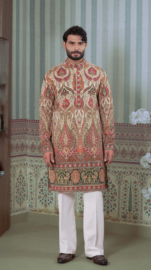 A man modeling a traditional Indian kurta in a beige, red, and green patterned design, paired with white trousers and brown shoes, standing in front of a patterned wall.