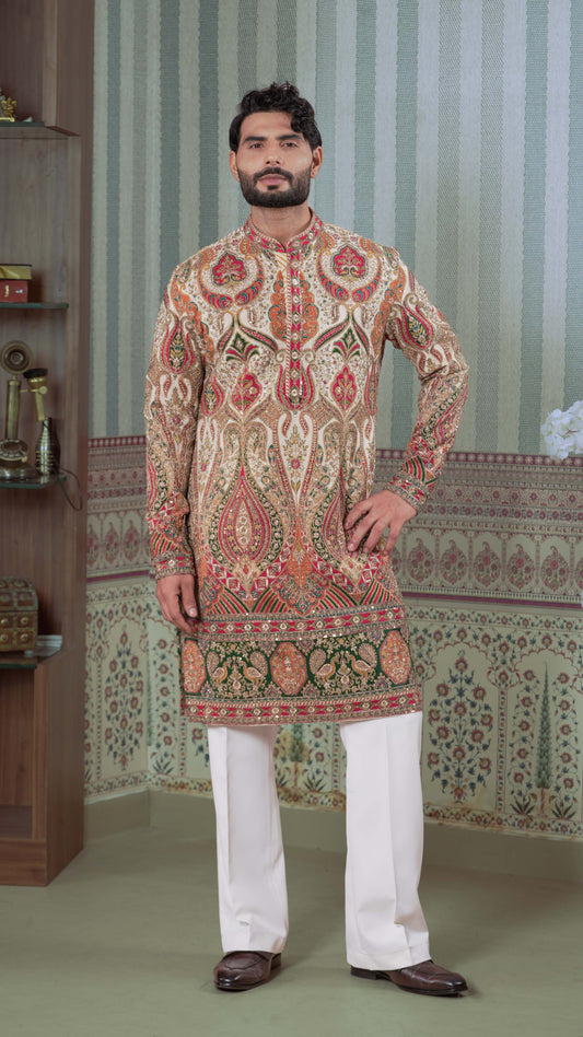 A man modeling a traditional long-sleeve kurta with an intricate multicolor paisley and floral pattern in beige, red, and green, paired with white trousers and brown shoes, standing in a decorated room with patterned wallpaper.