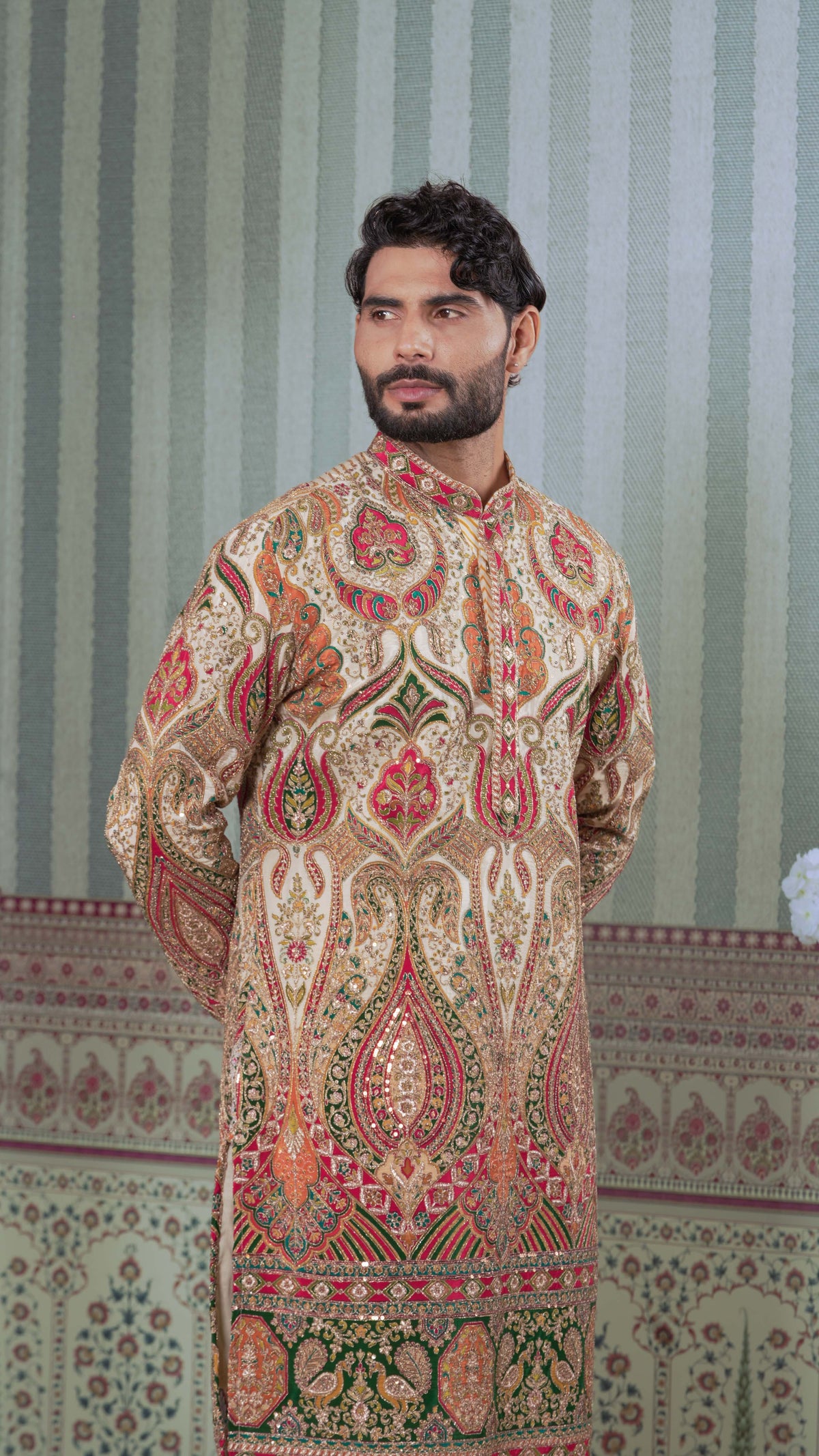 A man wearing a long-sleeved, high-collar traditional kurta with an intricate, ornate paisley and floral pattern in beige, red, green, and gold. He is standing in front of a patterned wallpaper background.