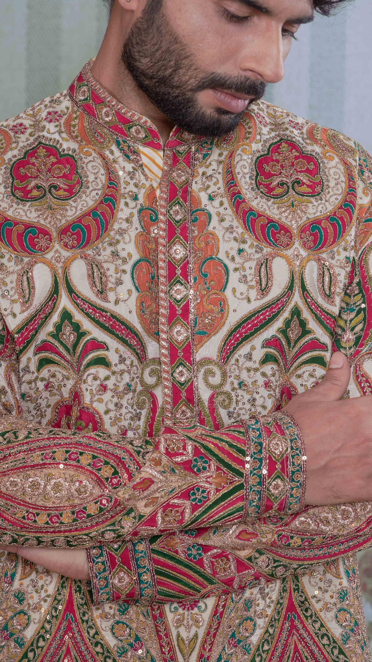 A close-up of a man wearing a heavily embroidered traditional kurta with intricate paisley and floral motifs in red, green, gold, and beige.