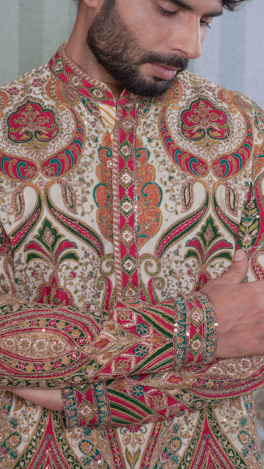 A close-up of a man wearing a heavily embroidered traditional kurta with intricate paisley and floral motifs in red, green, gold, and beige.