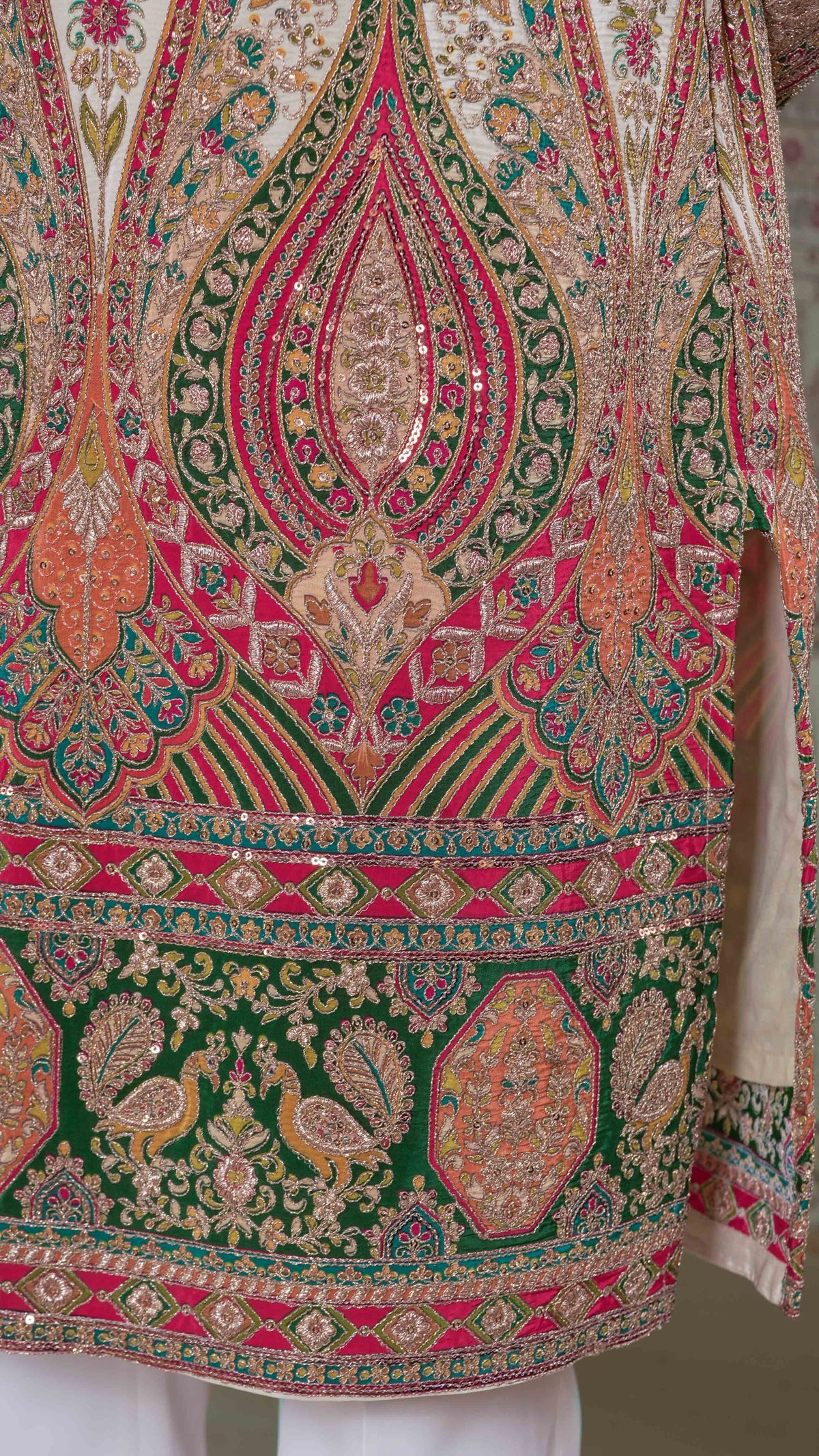 Close-up of an ornate embroidered tunic with intricate paisley and floral patterns in green, pink, orange, and gold, featuring peacock motifs and sequined detailing.