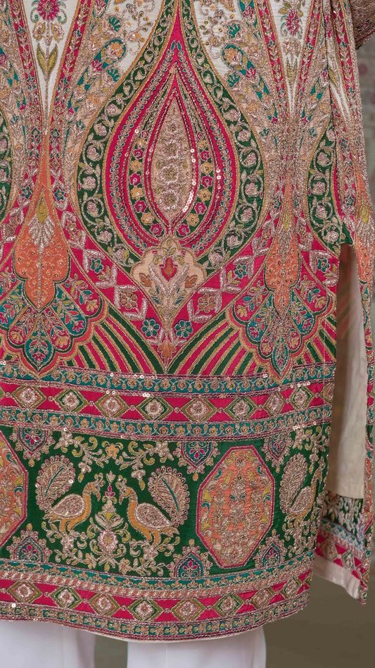 Close-up of an ornate embroidered tunic with intricate paisley and floral patterns in green, pink, orange, and gold, featuring peacock motifs and sequined detailing.